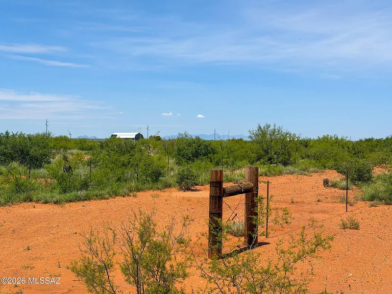Metal gate frame at a lot entrance on red dirt, with a small outbuilding and mesquite scrub beyond