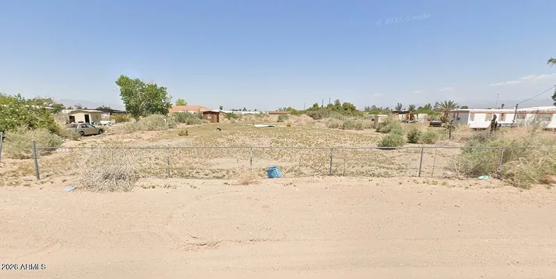 Street-level view of the flat sandy lot with a chain-link fence and a neighboring home behind